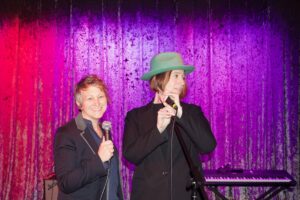 Rabbis Nicole Fix and Bronwen Mullion on stage, holding microphones, standing in front of a keyboard