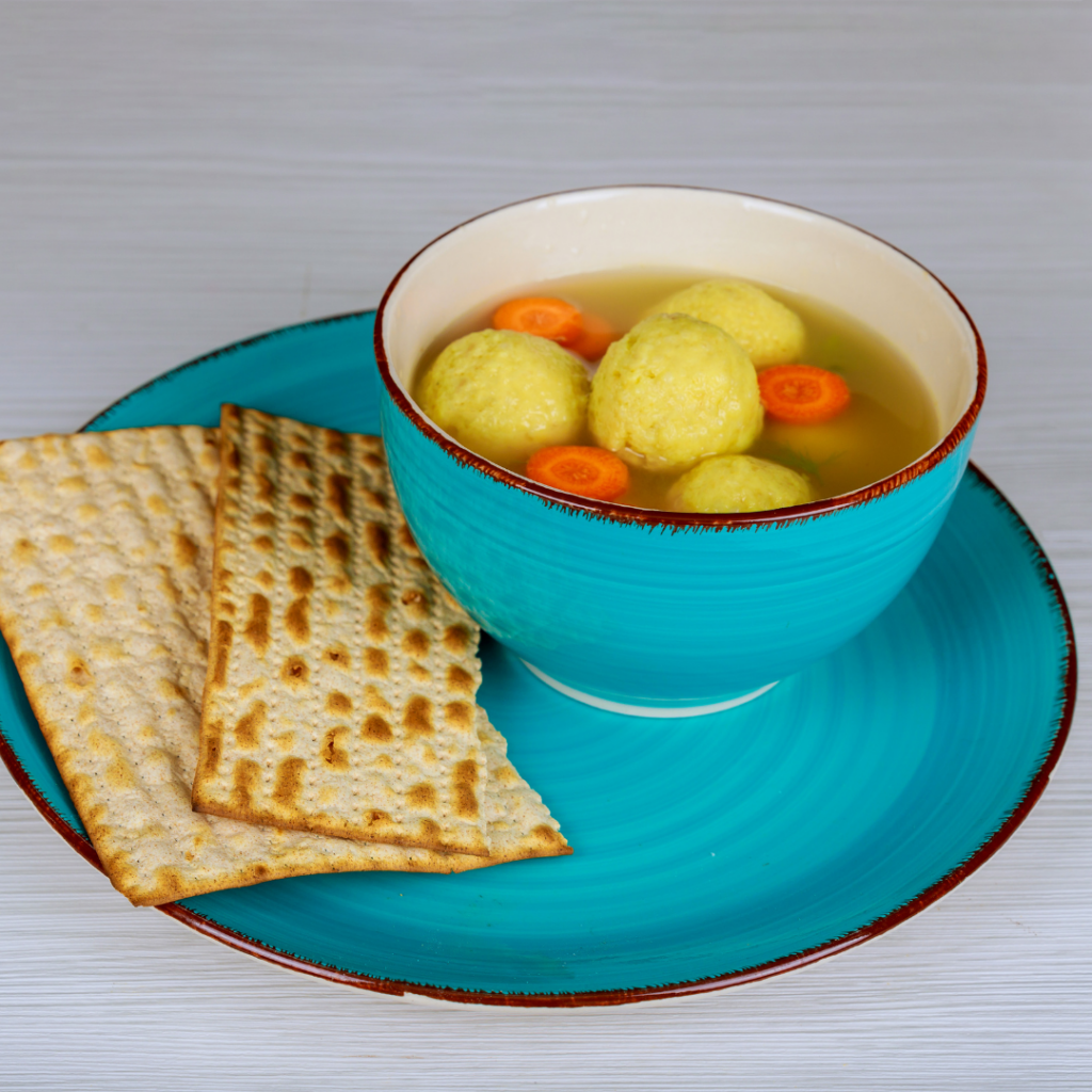 Blue bowl of matzo ball soup with carrot slices, served on a matching plate with matzo crackers.