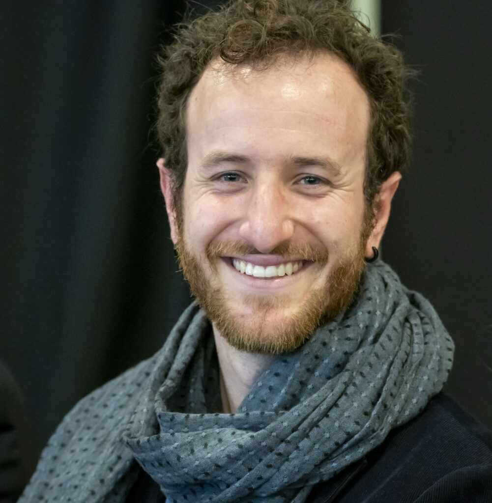 Smiling man with curly hair and a beard, wearing a gray scarf and dark clothing against a blurred background.