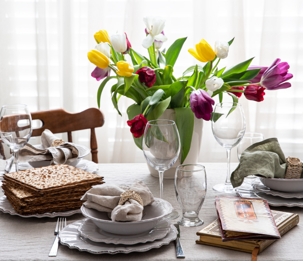 Elegant Passover table setting with matzos, tulips, glasses, and books against sheer curtains.