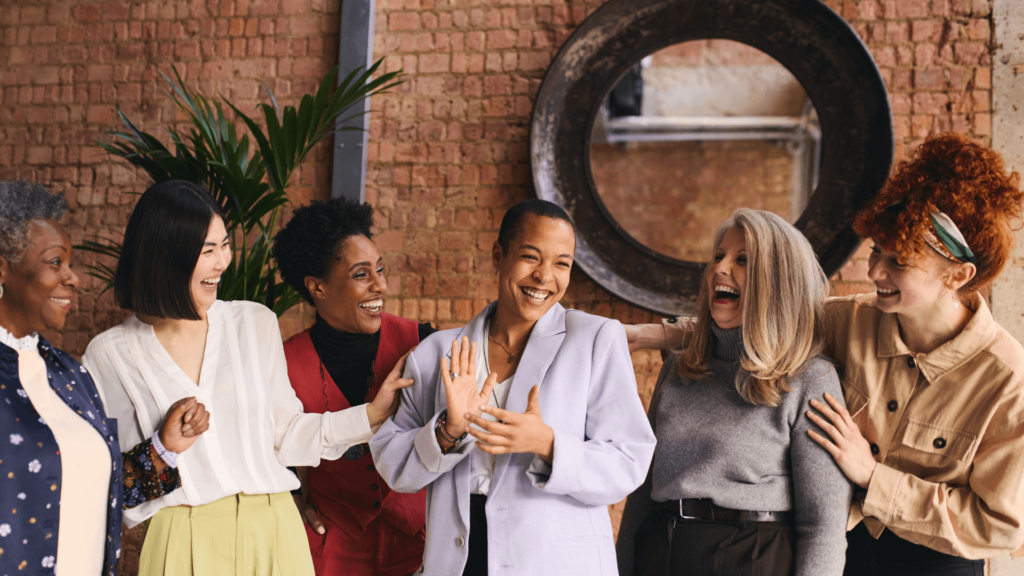 A multicultural group of women talking and laughing indoors against an exposed brick wall