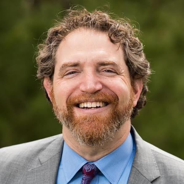 Smiling man with curly hair and beard, wearing a suit and tie, in front of a blurred green background.
