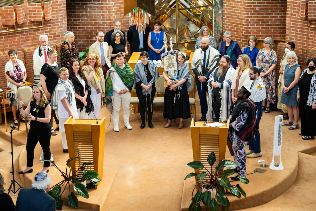 A diverse group of people gathers in a synagogue, some holding a Torah scroll, in a brightly lit room.