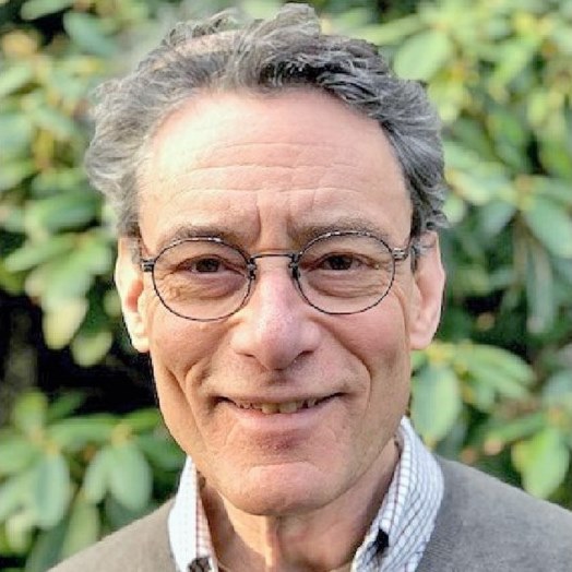 An older man with glasses smiles, standing in front of green foliage.