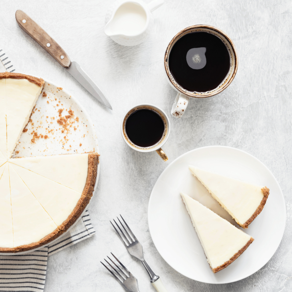 Top view of a cheesecake with slices on a plate, two coffee cups, forks, and a knife on a light surface.