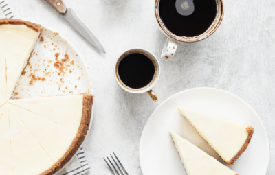 Top view of a cheesecake with slices on a plate, two coffee cups, forks, and a knife on a light surface.