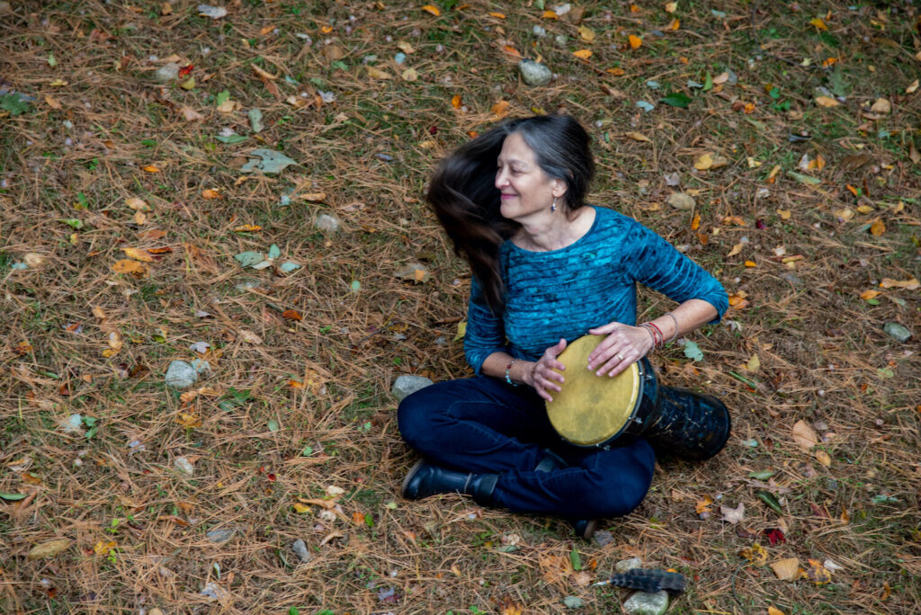 A person sitting cross-legged on the ground playing a drum, surrounded by fallen leaves.