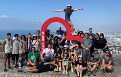 Group of people posing around a Love Haifa sign with a scenic view of the city and sea in the background.