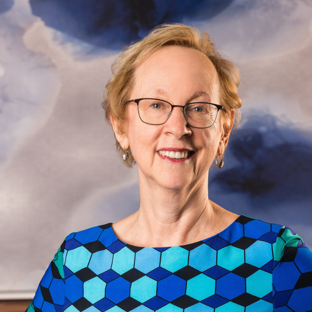 Smiling woman with glasses in a blue geometric-patterned shirt, standing against an abstract blue and white background.