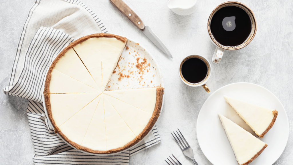 A sliced cheesecake on a plate next to a smaller plate with cake slices and cups of coffee