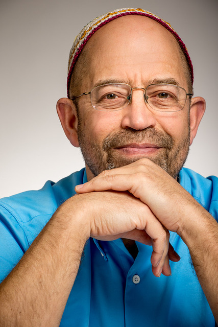 A smiling man with glasses and a colorful cap, resting his chin on his hands, wearing a blue shirt.