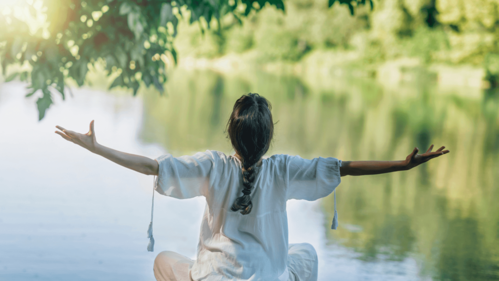 A woman sitting at the edge of a lake with her arms outstretched