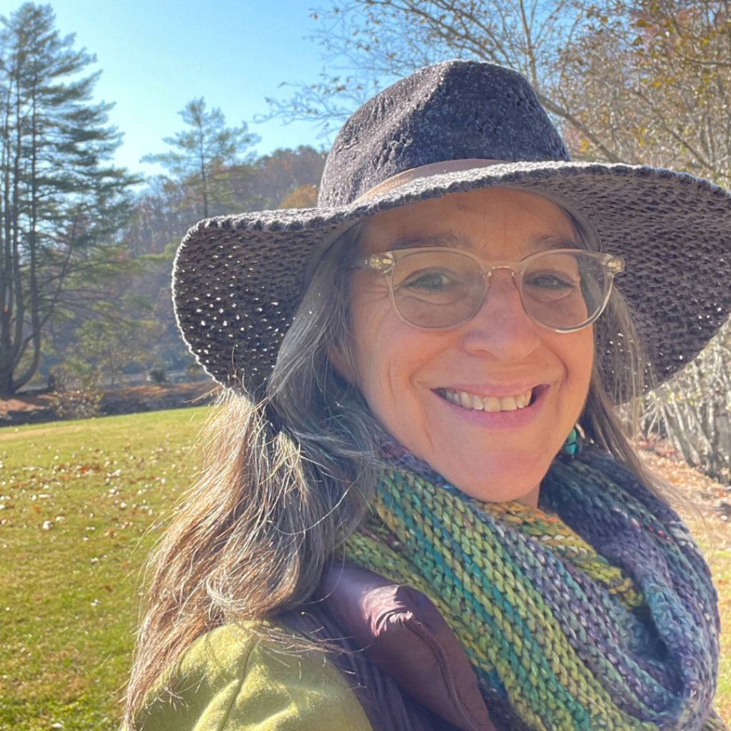 A woman wearing a hat and scarf smiles while standing outdoors in a sunny, leafy setting.