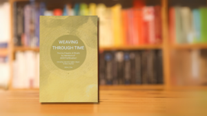Book titled Weaving Through Time on a wooden table, blurred colorful bookshelf in the background.