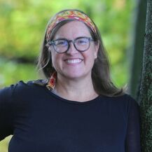 Smiling person with glasses and a colorful headband stands near a tree in a green, blurred outdoor setting.
