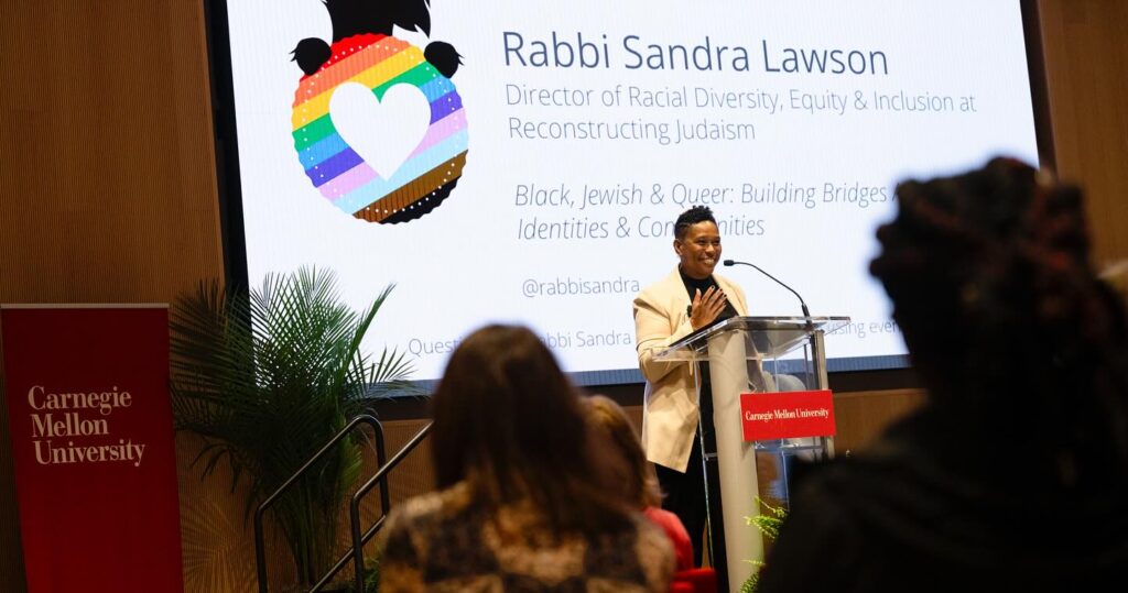 Rabbi Sandra Lawson speaks at Carnegie Mellon University event, standing at a podium with a projected slide behind her.