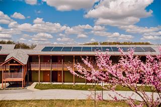 Building with solar panels on the roof, pink flowering tree in the foreground, partly cloudy sky.