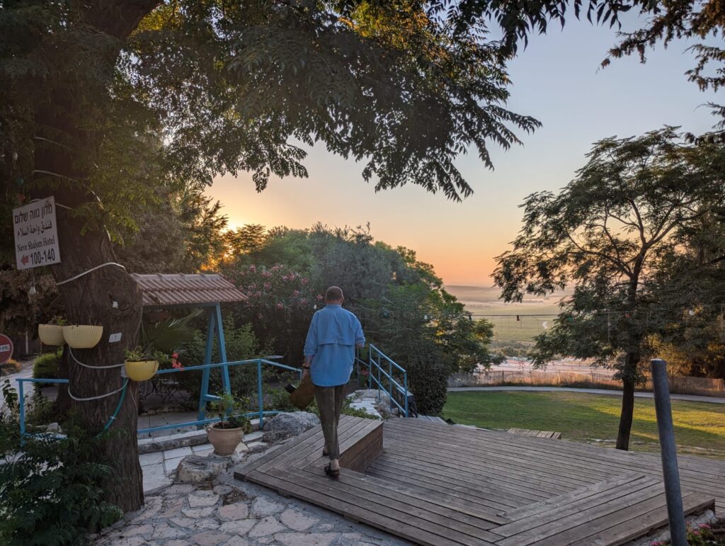Person walking on wooden deck towards a beach at sunset, surrounded by trees and a garden.