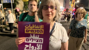 A woman holds a multilingual protest sign during a nighttime street demonstration.