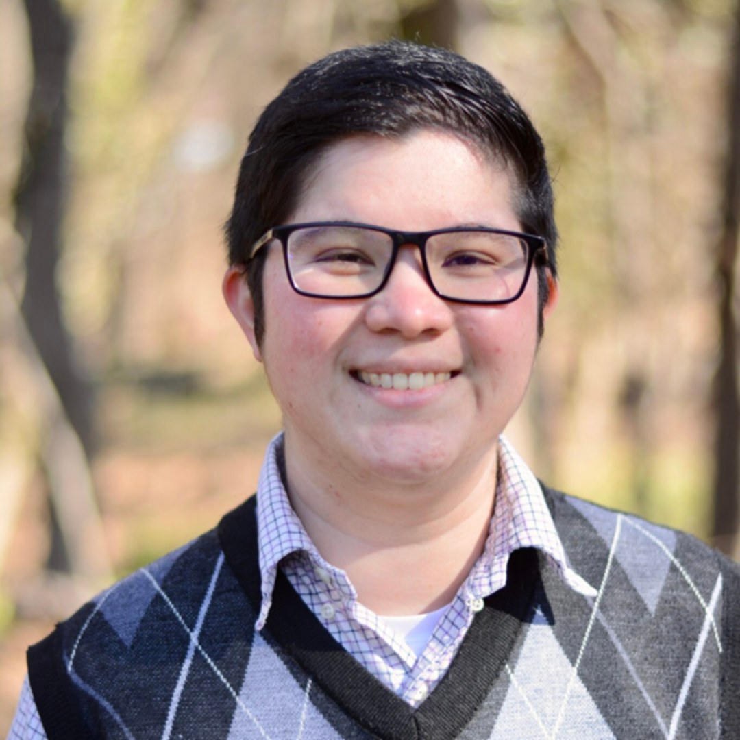 A person wearing glasses and a sweater vest smiles outdoors with trees in the background.