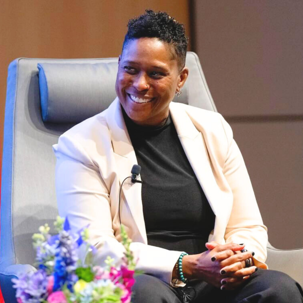 Person smiling while sitting in a chair, wearing a light blazer and black top, with flowers in the foreground.