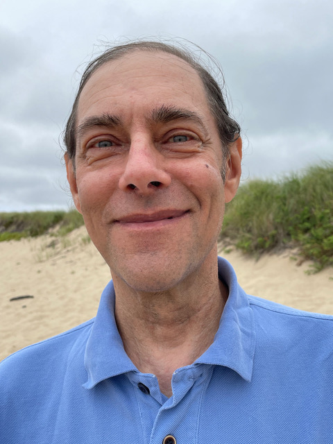 Man in a blue polo shirt smiles at the beach, with grassy dunes in the background under a cloudy sky.