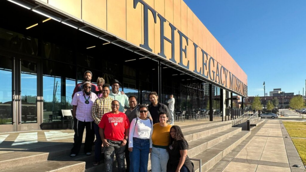 A diverse group poses in front of The Legacy Museum building on a sunny day.
