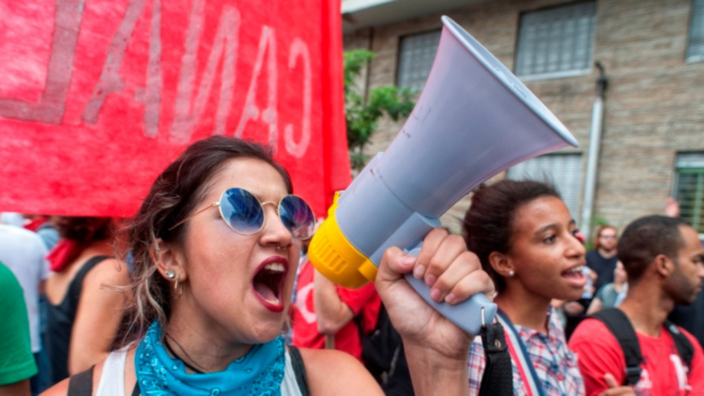 Protesters with a megaphone and a red banner, one person wearing sunglasses and a blue bandana.