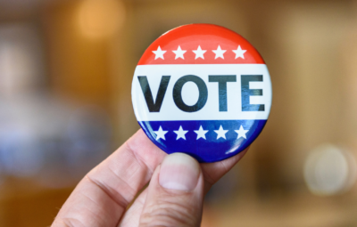 A hand holds a red, white, and blue button with the word VOTE and stars.