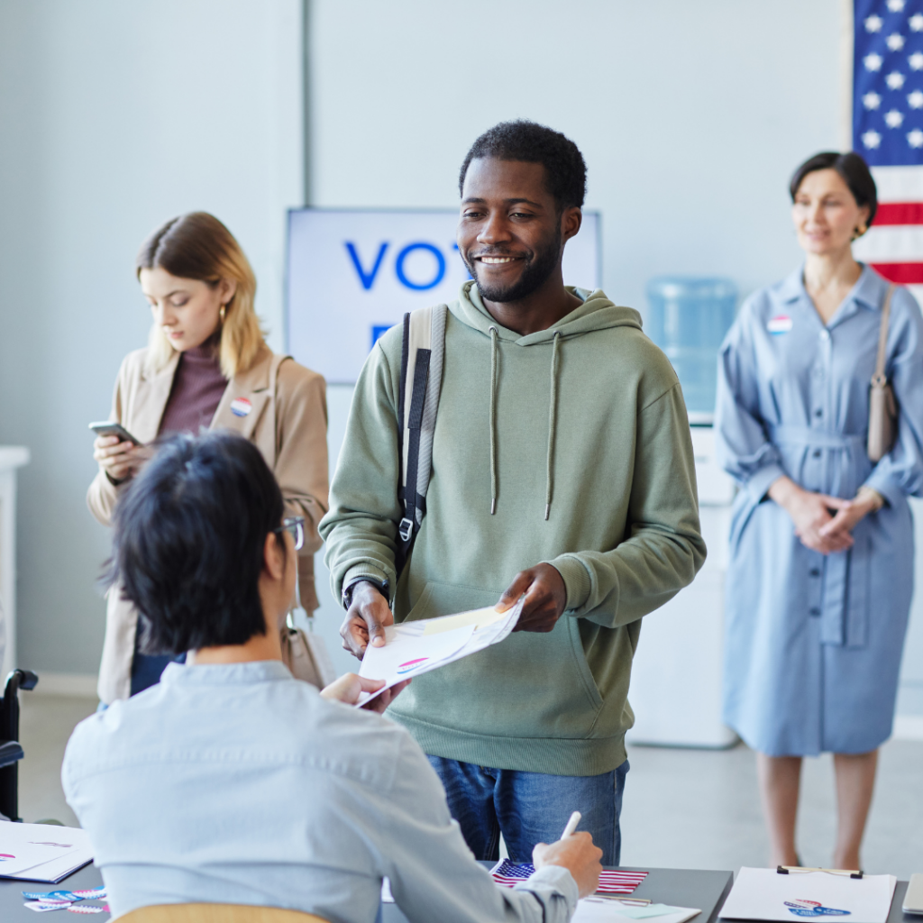 People at a voting station, one person handing a document to an official, others waiting in line.