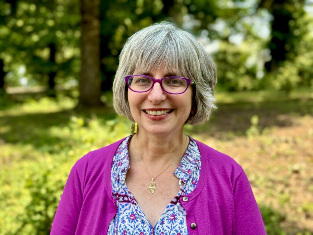 Smiling person with gray hair and glasses, wearing a pink cardigan, stands in a wooded area.