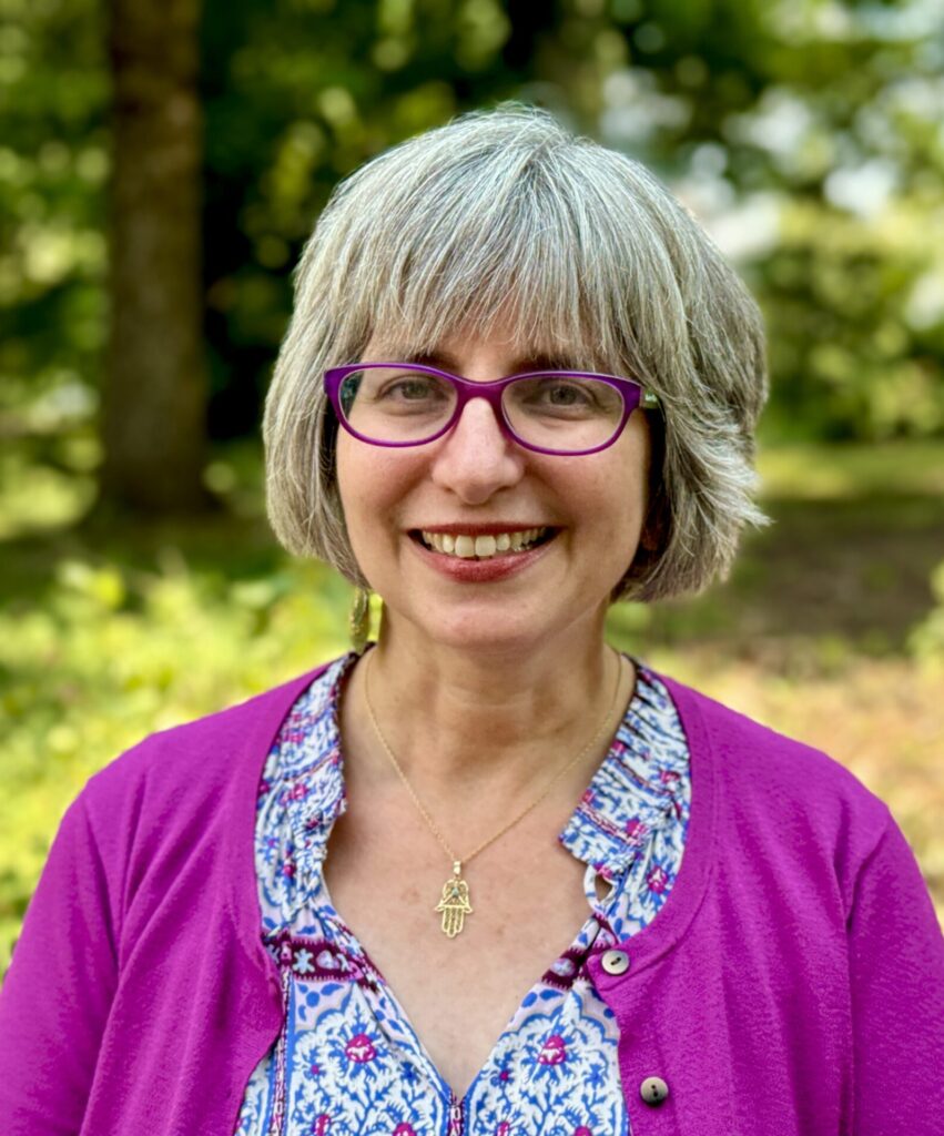Smiling woman with gray hair, wearing purple glasses and a pink cardigan, standing outdoors with greenery in the background.