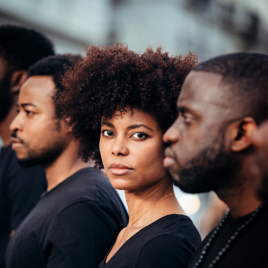 A woman with curly hair looks at the camera, standing among three men in the street.