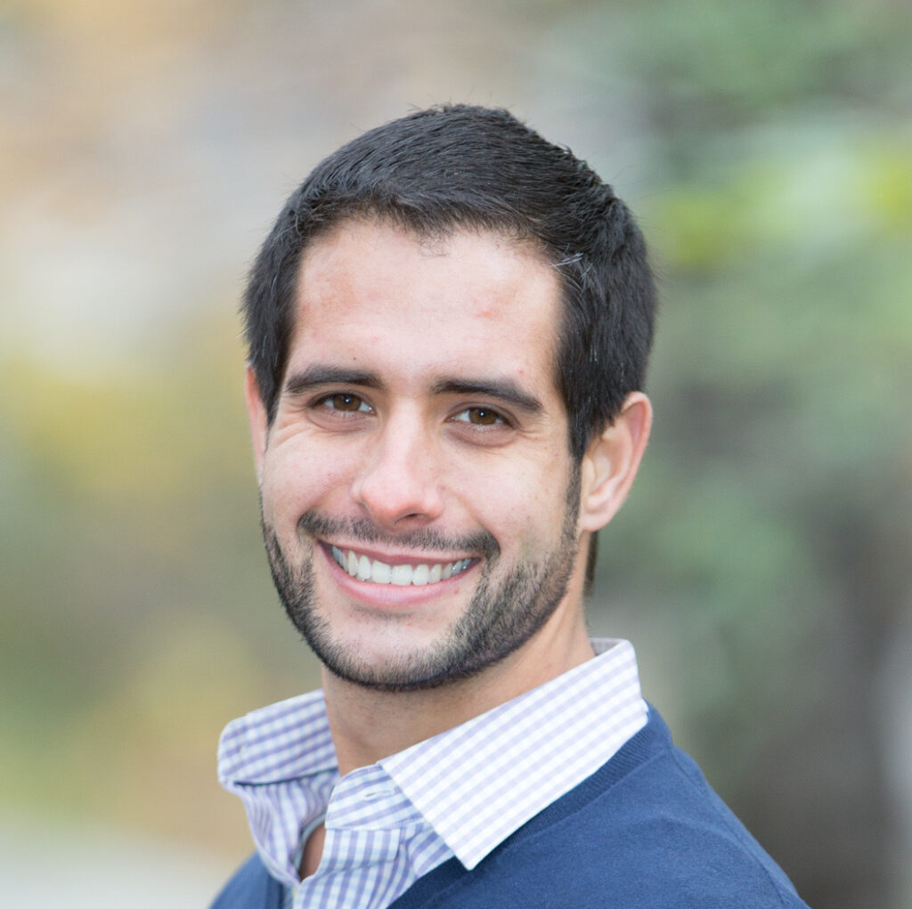 Justin Rosen Smolen smiles in front of a blurred outdoor background. He's wearing a blue sweater and a dress shirt with the top button undone.