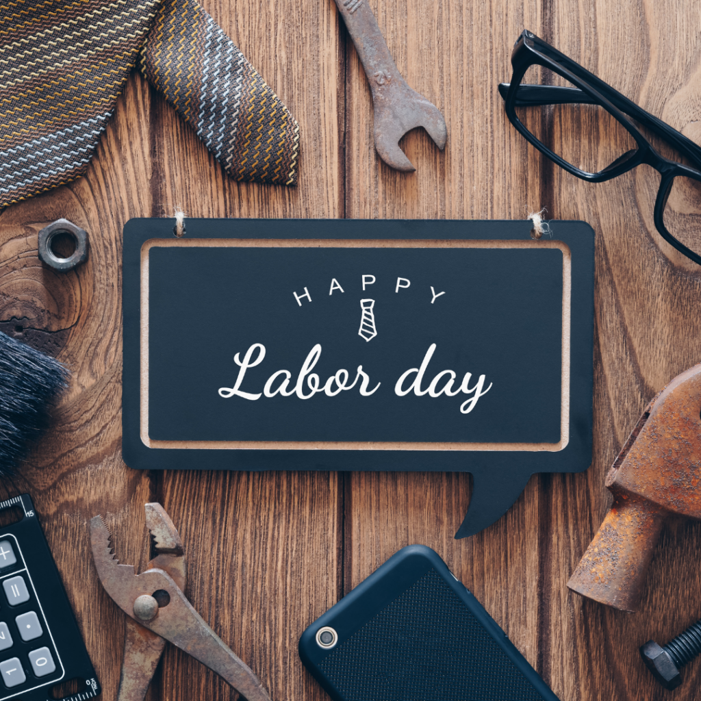 A Happy Labor Day sign on a wooden desk surrounded by tools, glasses, and neckties.