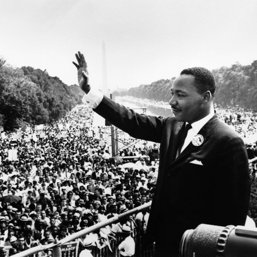 Man in a suit waves to a large crowd on a sunny day, with trees and a tall monument in the background.