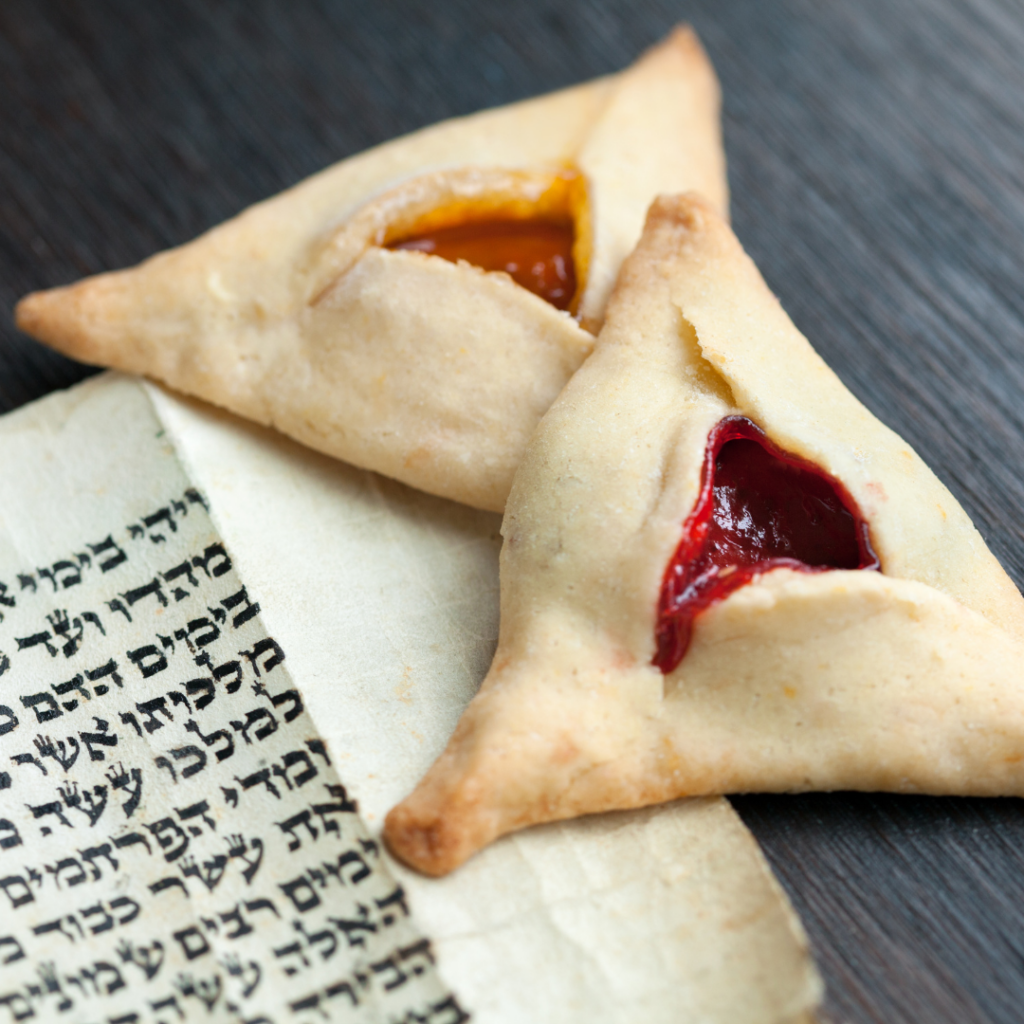 Two hamantaschen cookies with fruit fillings next to a Hebrew text scroll on a dark surface.