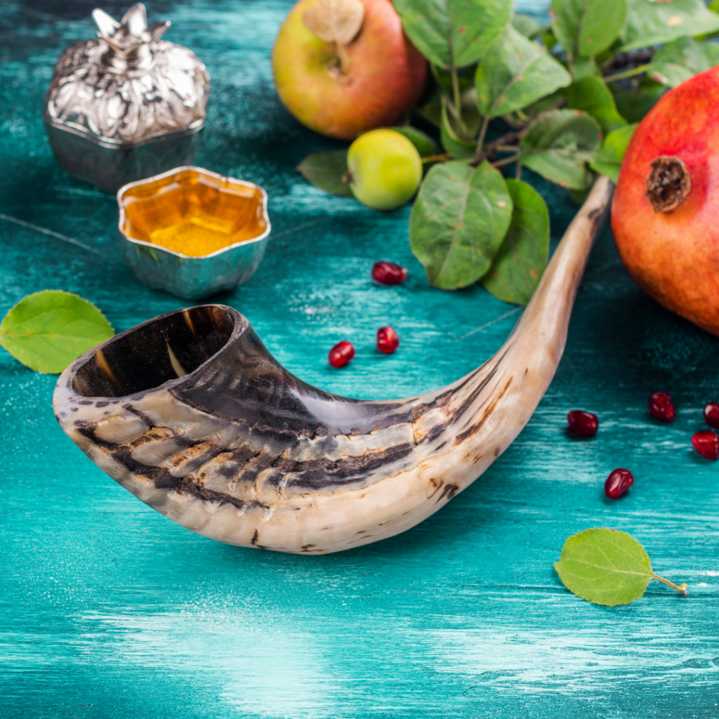 Shofar on a turquoise table with apples, pomegranate, leaves, and a small dish of honey.