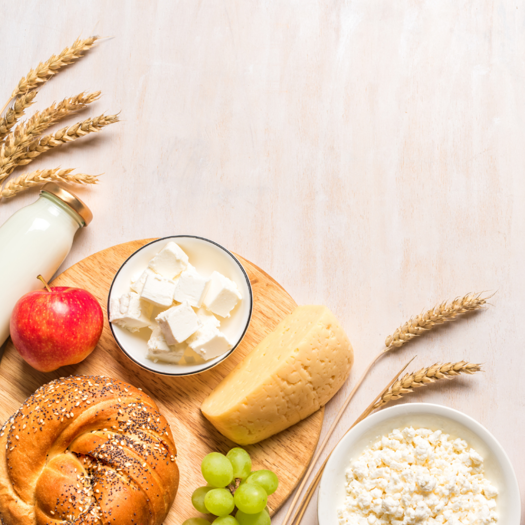 Breakfast scene with cheese, apple, grapes, yogurt, and a bagel on a wooden board, next to a milk bottle and wheat.