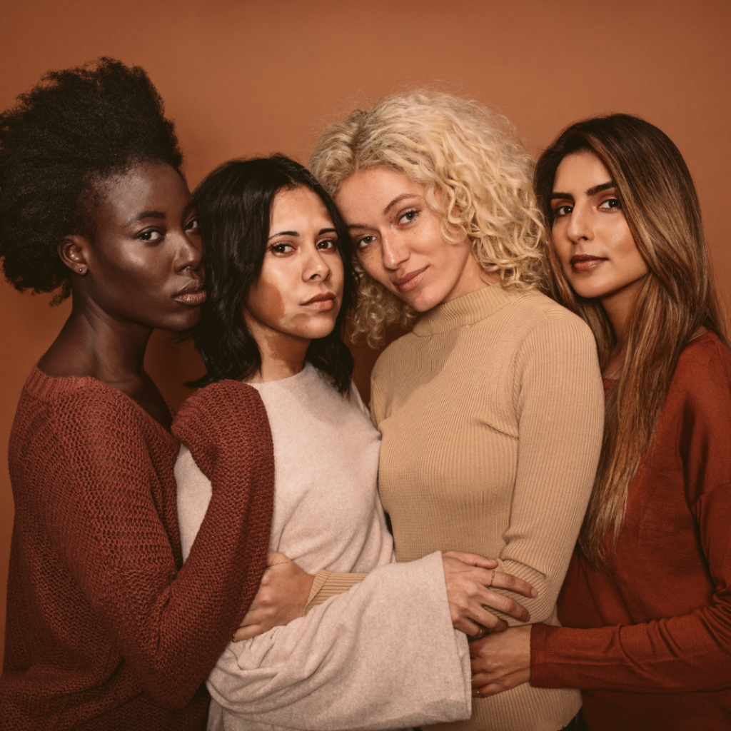 Four women of diverse backgrounds pose together against a brown backdrop, embracing each other closely.