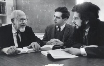 Three men in discussion at a table with open books, Hebrew text on the blackboard behind them.