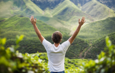 Person in white shirt standing with arms raised, facing lush green hills.