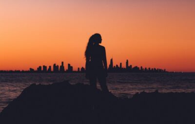 Silhouette of a woman standing on rocks, with a city skyline and orange sunset in the background.
