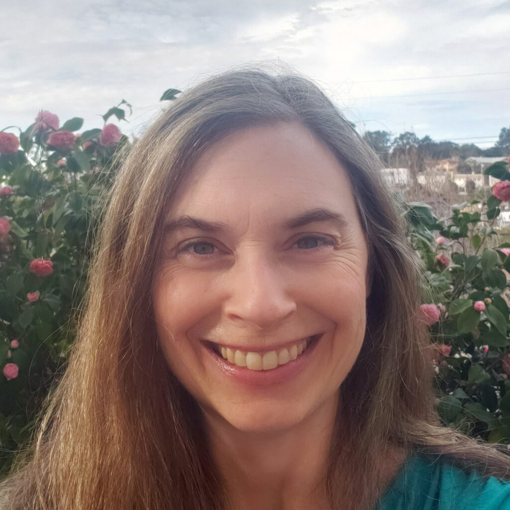 A woman smiling in front of a garden with pink flowers.