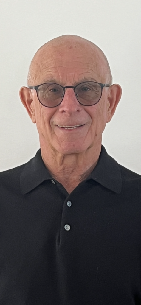 Elderly man wearing glasses and a black polo shirt, smiling in front of a plain white background.