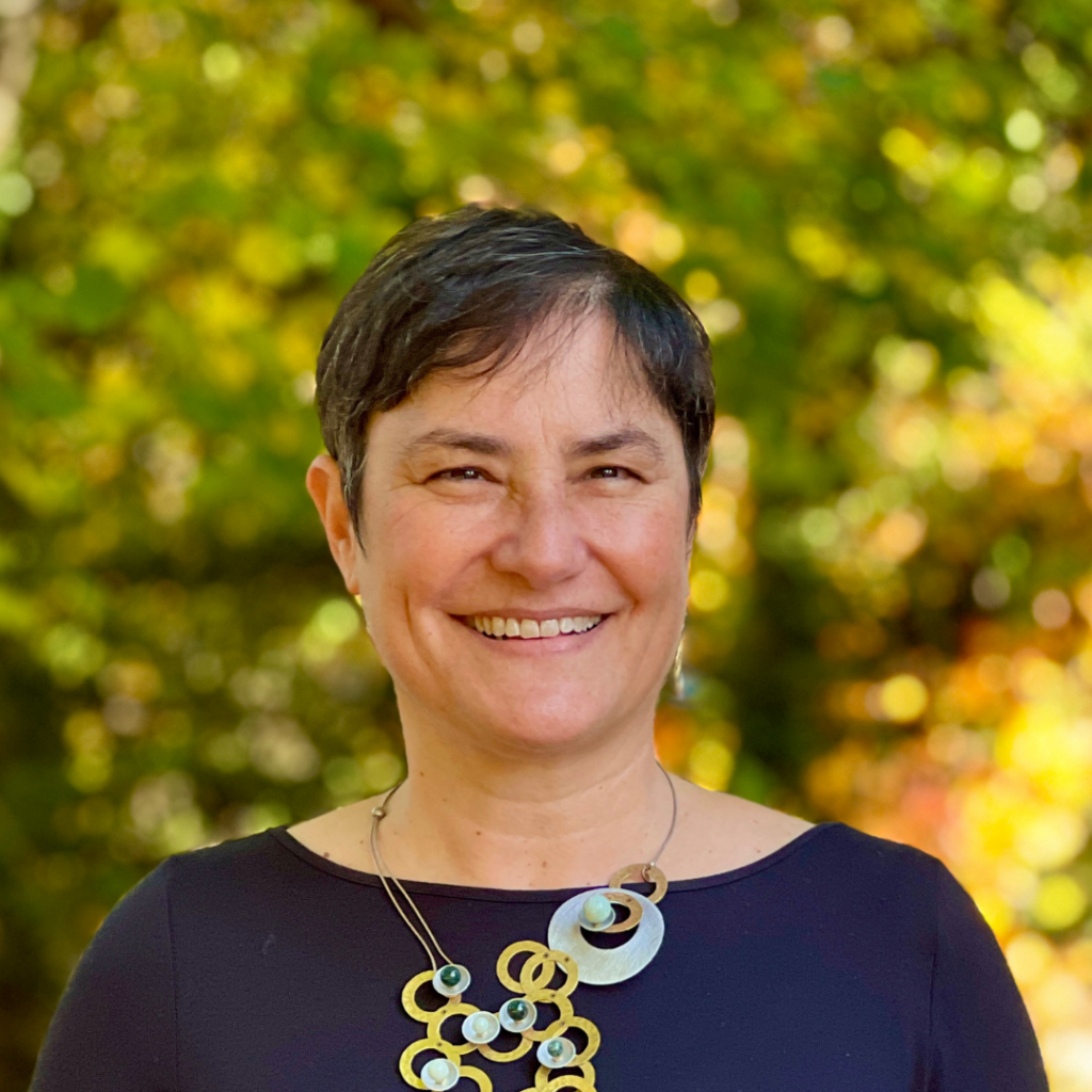 Smiling person with short hair and a necklace, standing outside with a blurred green and yellow leafy background.