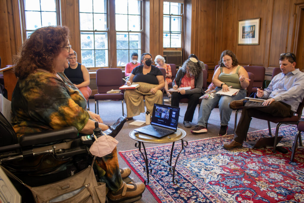 Wendy Elliott-Vandivier is seated, addressing a row of staff and students at the Reconstructionist Rabbinical College. Light pours in through the windows, illuminating a red floor rug.