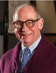 Elderly man wearing glasses, a maroon sweater, and a blue polka-dotted tie, smiling at the camera.