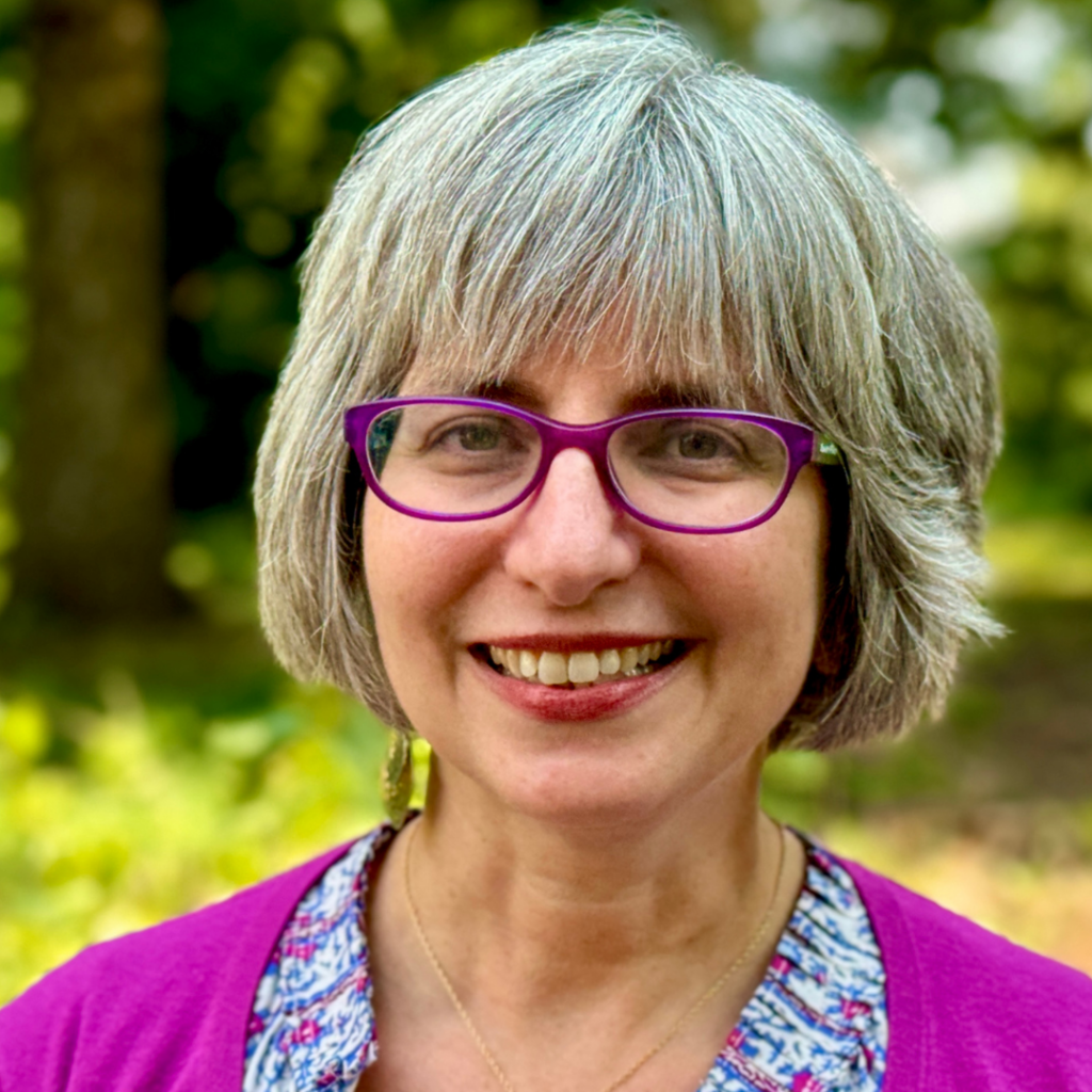 Rabbi Mira Wasserman, with gray hair and glasses, wearing a purple top, stands outside with greenery in the background.