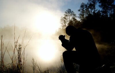 Silhouette of a person kneeling and praying by a misty lake with trees in the background at sunrise.